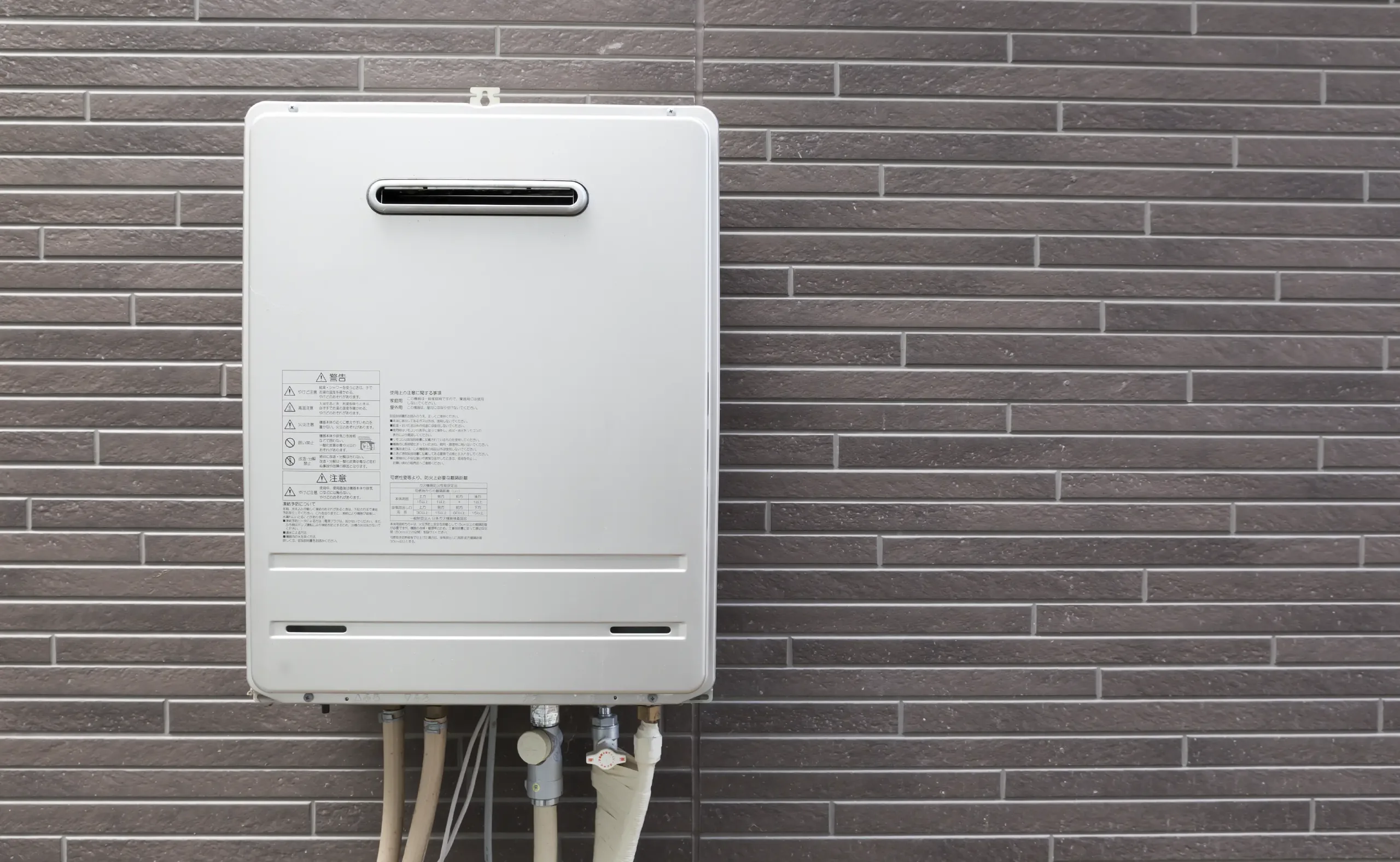 Tankless water heater mounted on an exterior brick wall with connected pipes, used for heating household water.