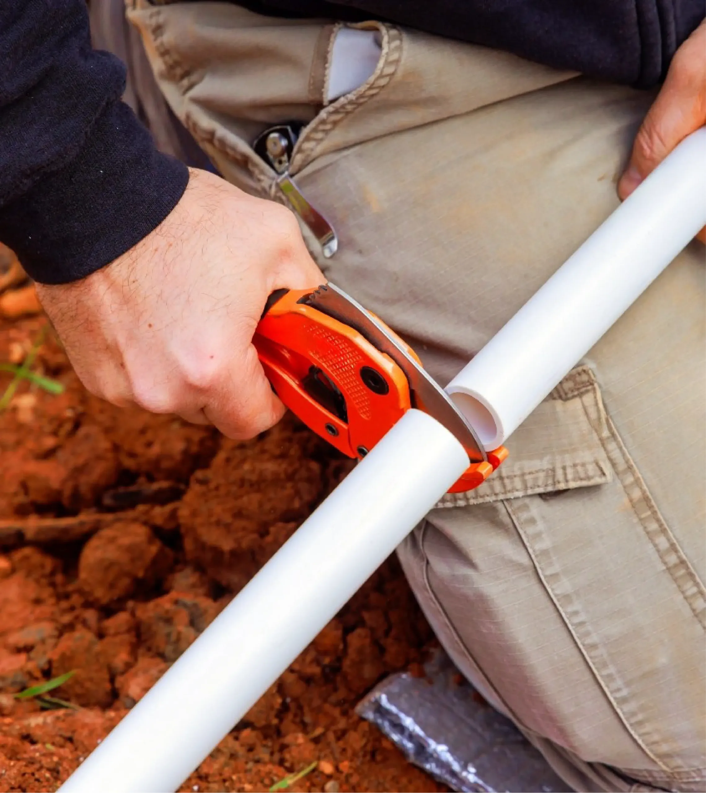 A person uses a pipe cutter to cut a white PVC pipe outdoors on the ground during a water line repair.