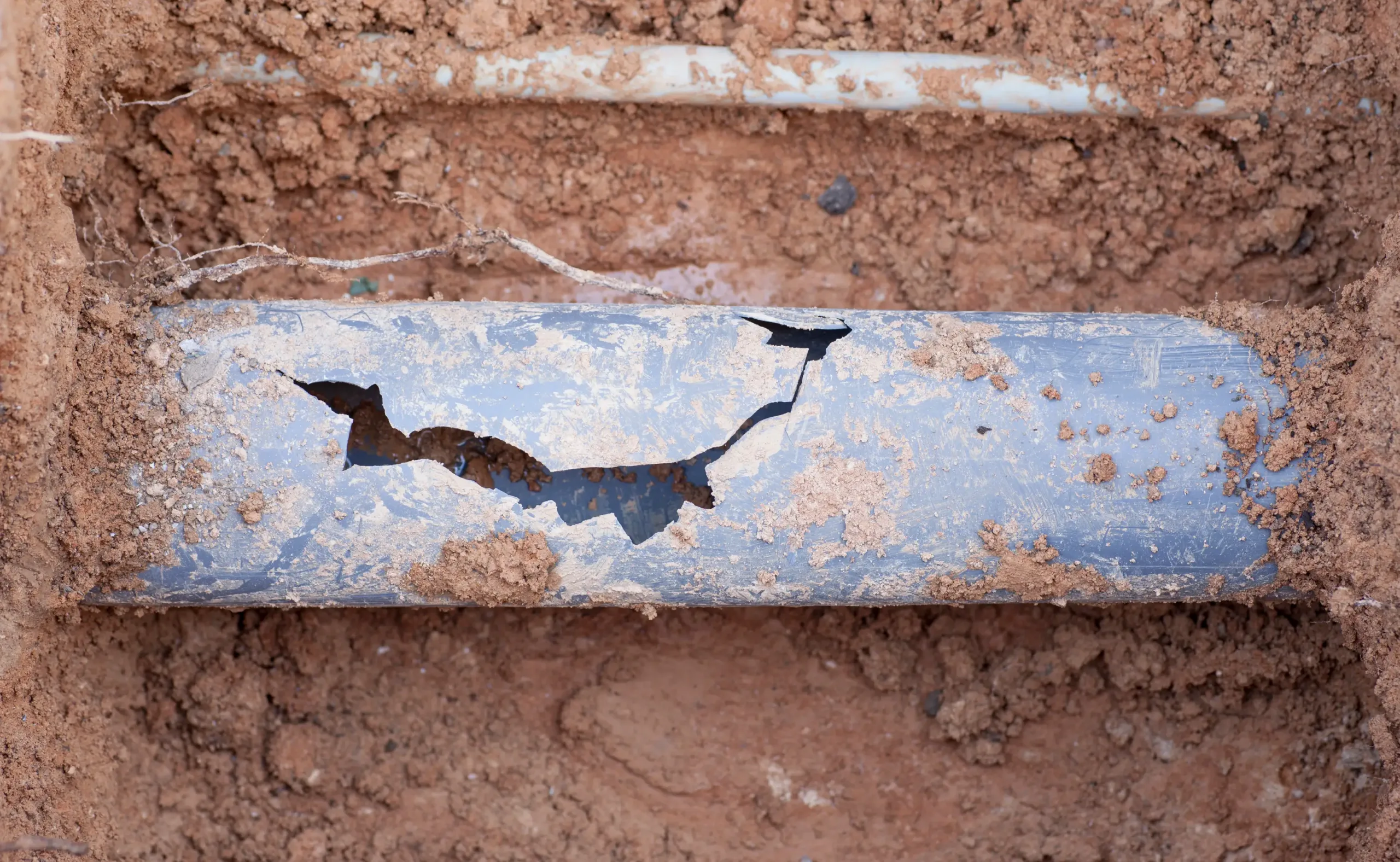 A close-up view of a damaged sewer line metal pipe with a large crack, partially covered in soil.