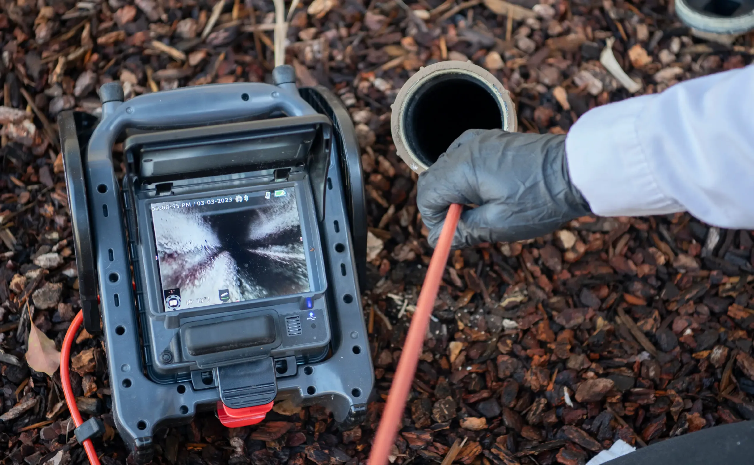 A gloved hand inserts a camera cable into a pipe for Sewer Camera Inspections, while a monitor displays the inside of the pipe surrounded by mulch.
