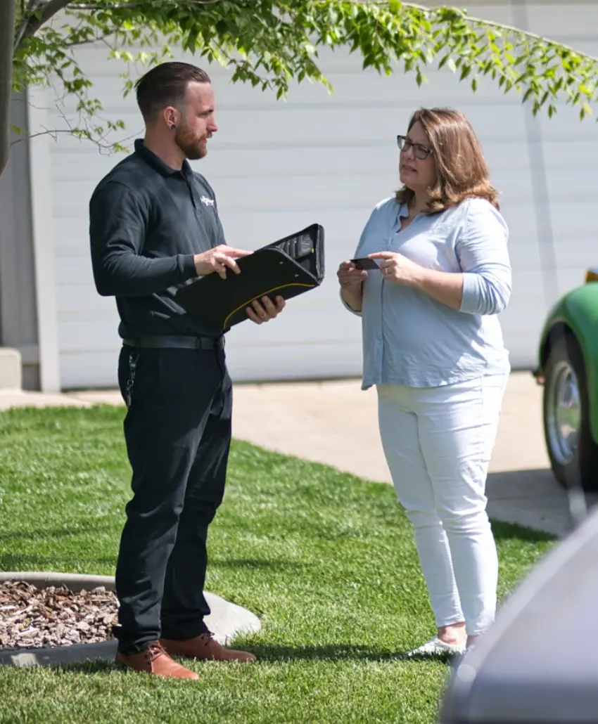 A man holding a folder speaks with a woman holding a card while standing on a lawn near a driveway and parked cars.