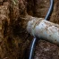 A close-up of a blue pipe partially buried in soil inside a trench, with a black cable running parallel to it and tree roots weaving through the earth nearby.